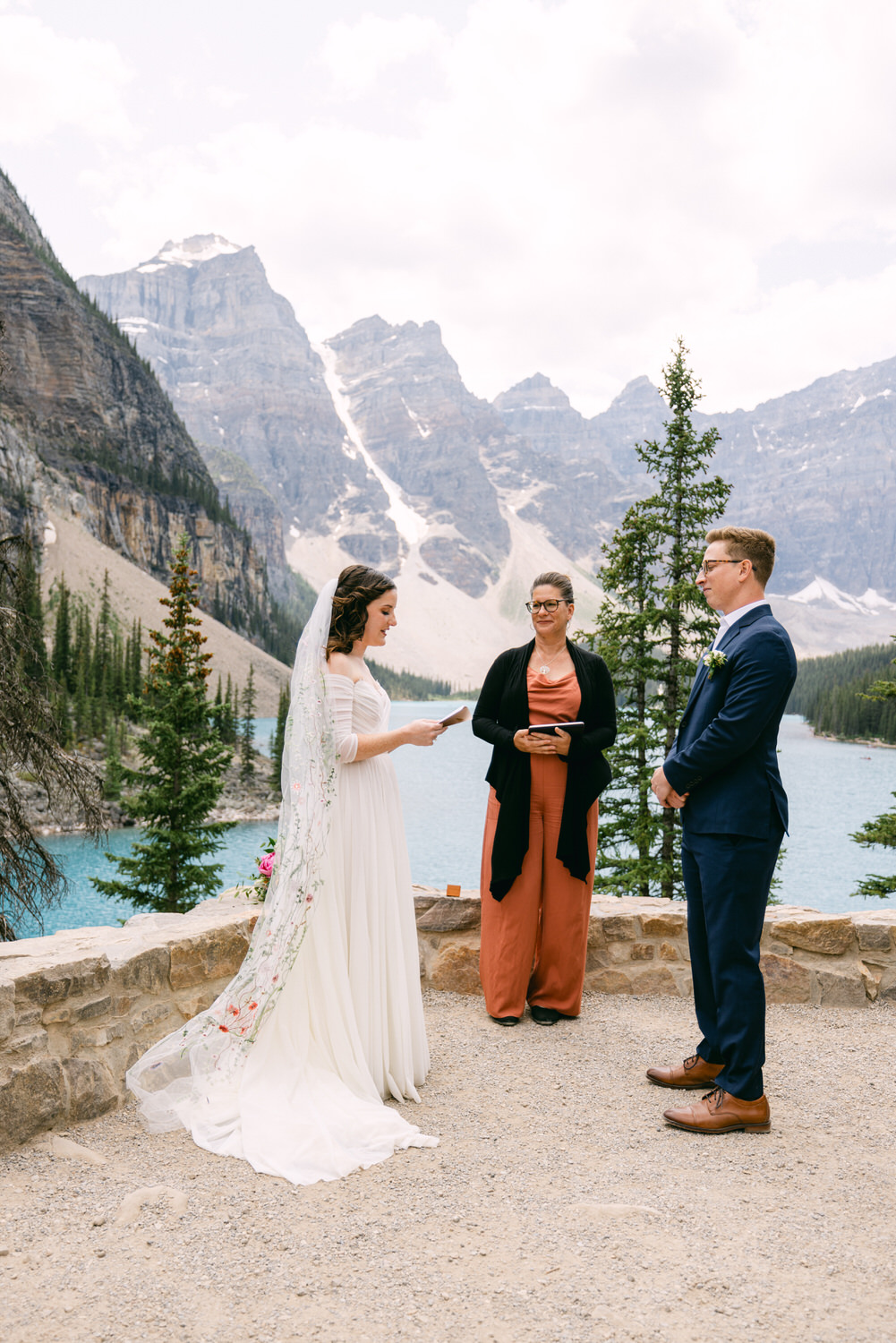 A bride and groom exchange vows during a scenic outdoor wedding ceremony by a lake surrounded by mountains and trees.