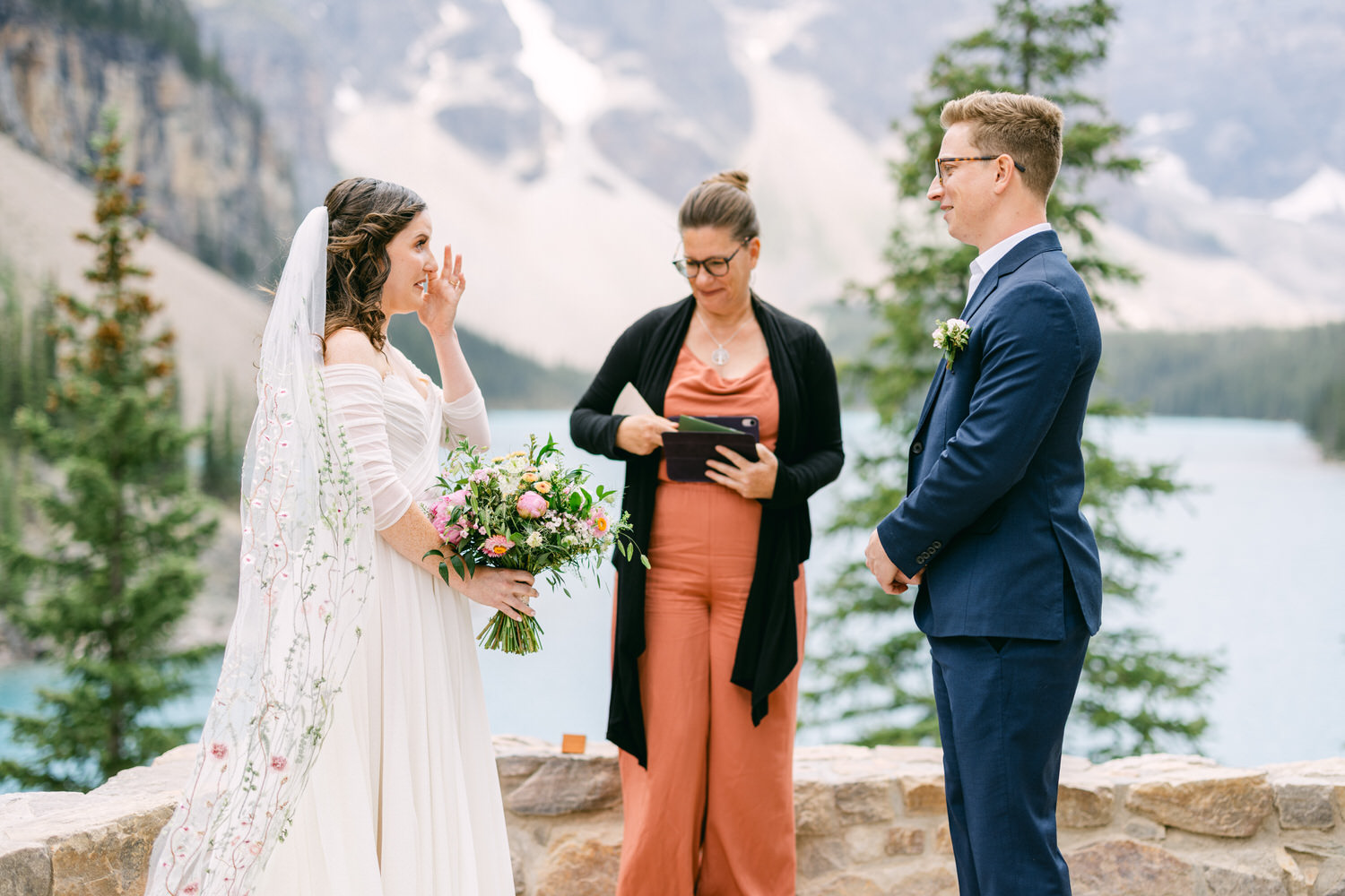 A couple exchanges vows during an outdoor wedding ceremony at a picturesque lakeside location, surrounded by mountains and trees. The bride, holding a bouquet, wipes away a tear while the officiant stands nearby.