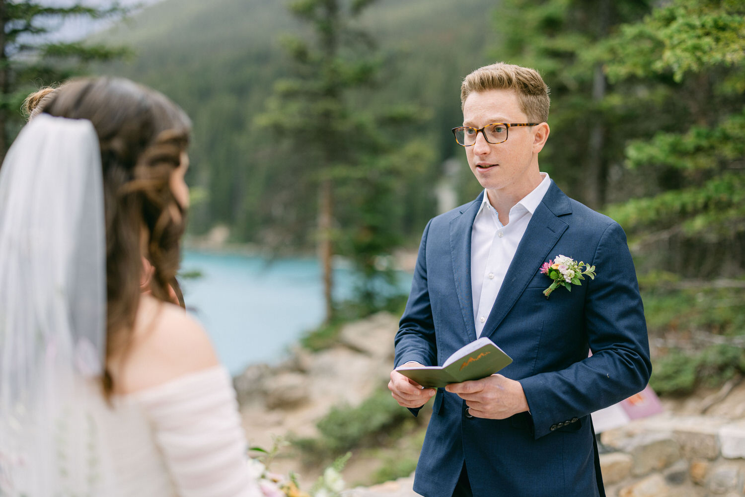 A groom stands speaking at an outdoor wedding ceremony, with a bride partially visible in the foreground and a serene lake in the background surrounded by trees.