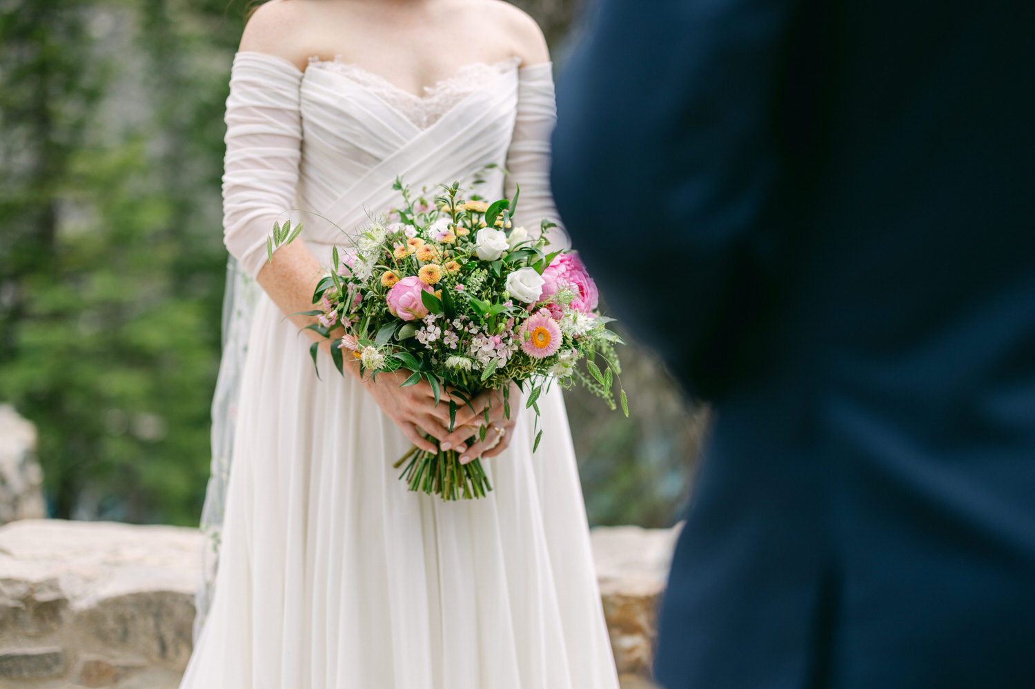 A bride holding a colorful bouquet of flowers, wearing a stunning white dress with off-shoulder sleeves, set against a lush green backdrop.