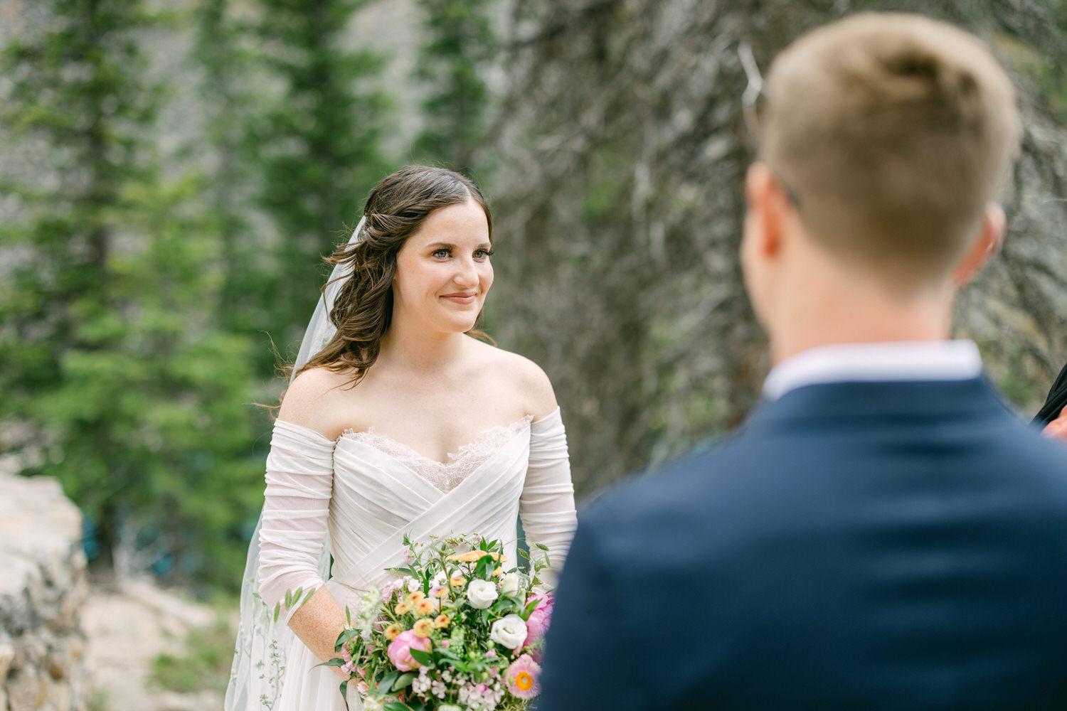 A smiling bride in a white wedding dress holds a colorful bouquet while engaging with her partner during a ceremony surrounded by trees.