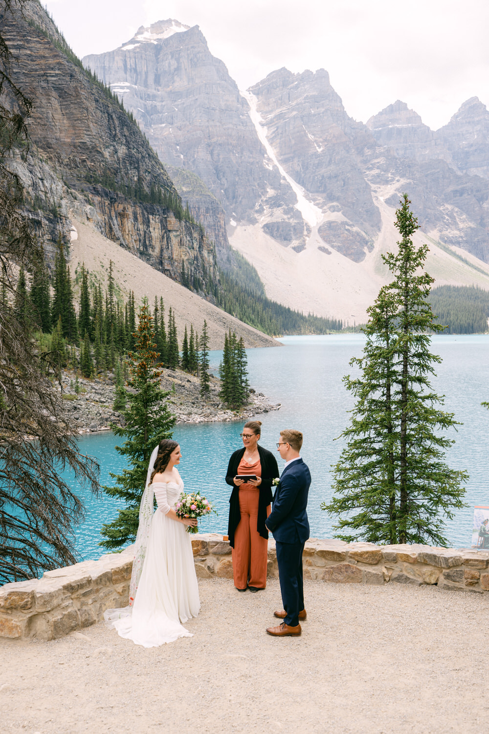 A couple exchanging vows during their wedding ceremony at a scenic lakeside location, surrounded by mountains and trees.