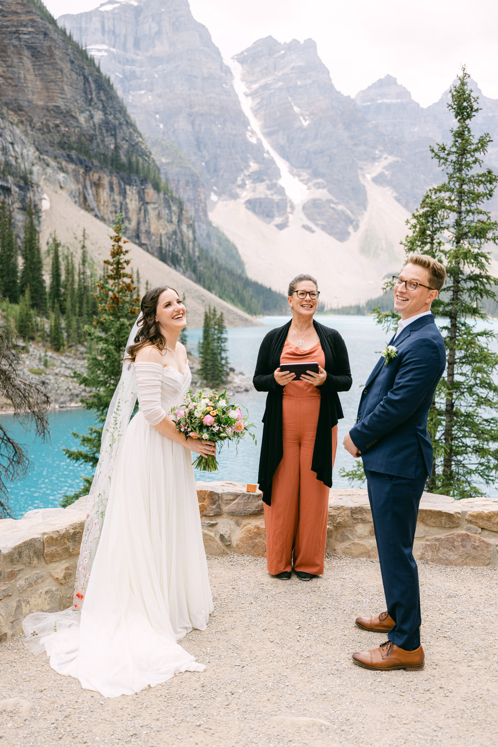 A joyful wedding couple stands by a serene lake surrounded by towering mountains, with an officiant smiling while leading the ceremony.