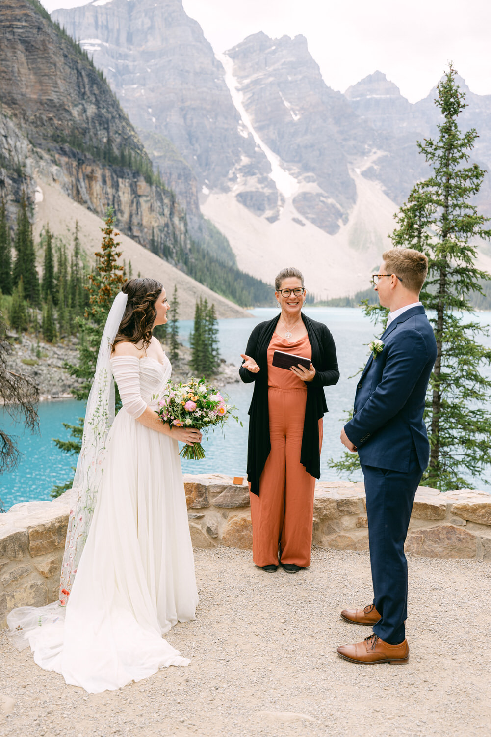 A couple exchanges vows during a scenic outdoor wedding ceremony by Moraine Lake, with mountains in the background and a celebrant officiating.