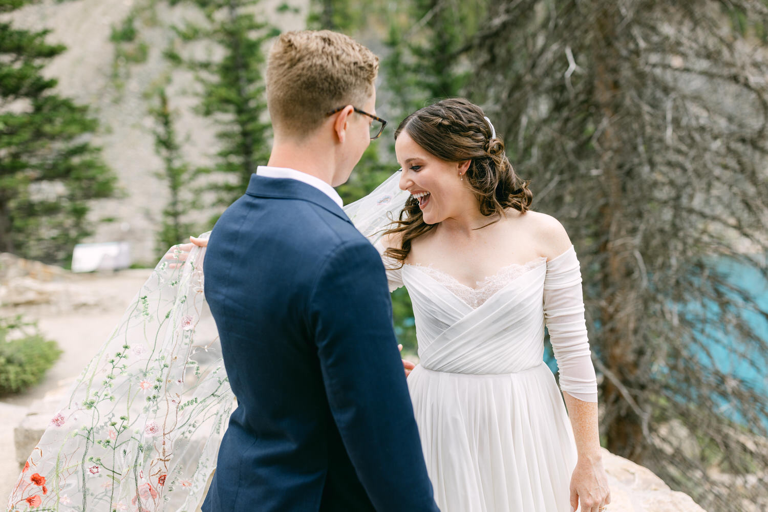 A couple shares a heartfelt laugh during their outdoor wedding ceremony, with the bride's flowing veil adorned with floral embroidery capturing the light.