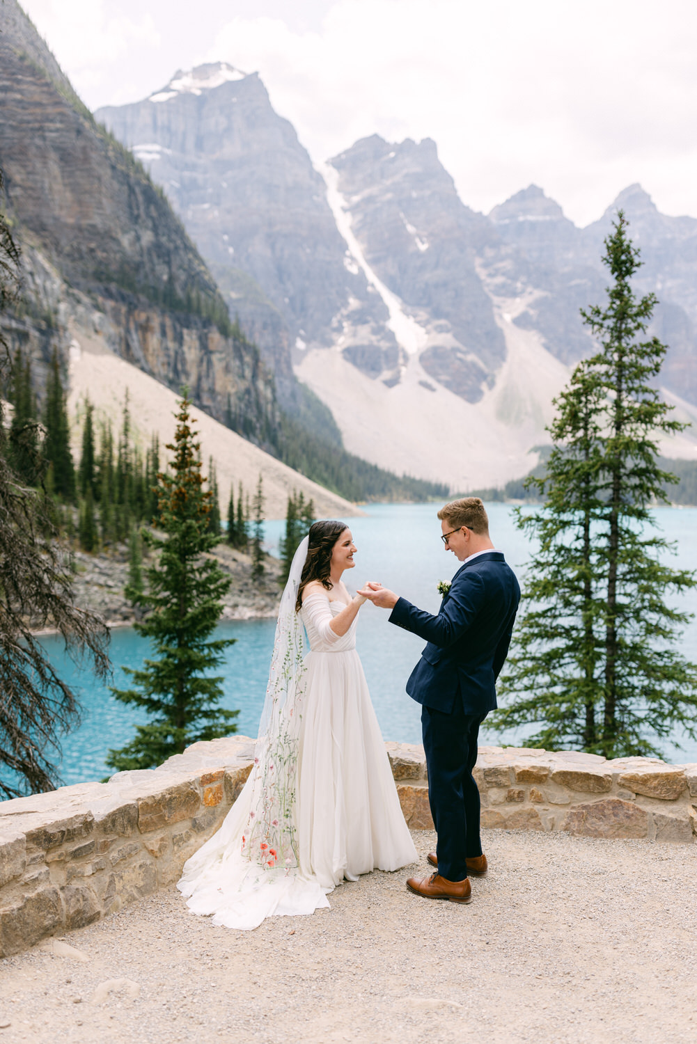 A couple exchanging vows against a stunning mountain backdrop with a turquoise lake, surrounded by lush trees.