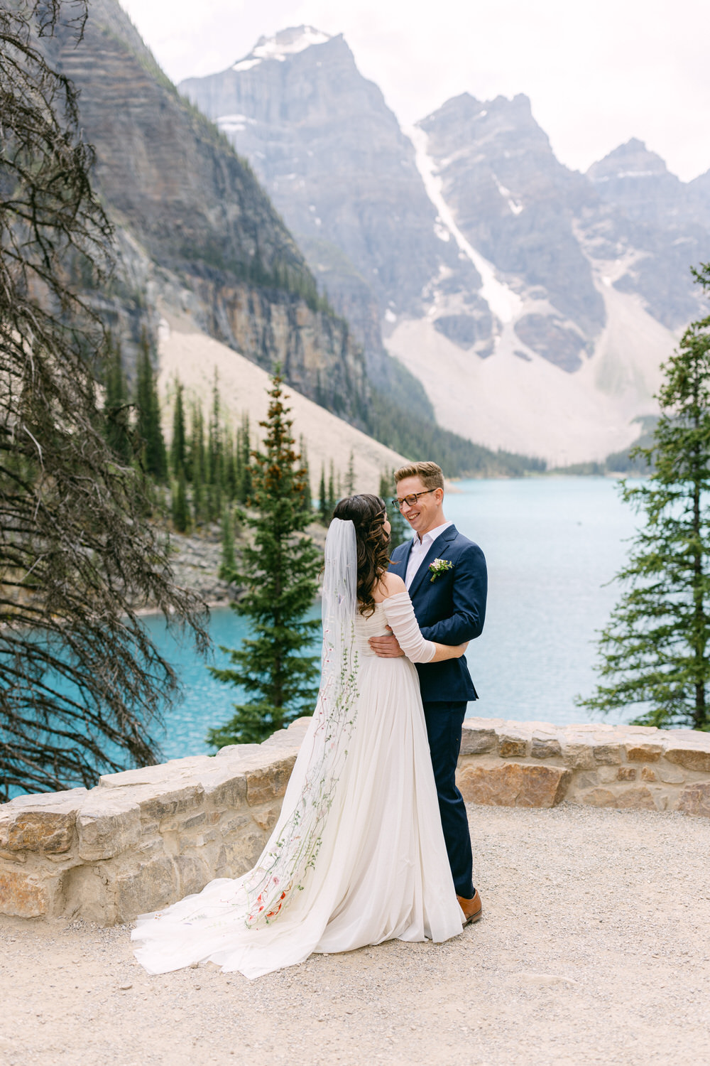 A couple embraces each other in a stunning outdoor setting, surrounded by majestic mountains and a serene blue lake, capturing a moment of love and joy.