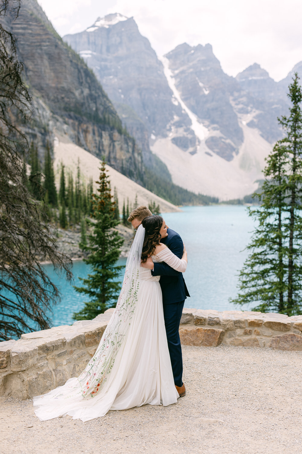 A joyful couple shares a romantic hug in a picturesque setting by a turquoise lake, surrounded by majestic mountains and lush greenery. The bride wears a flowing white gown with floral designs, while the groom is dressed in a navy suit.