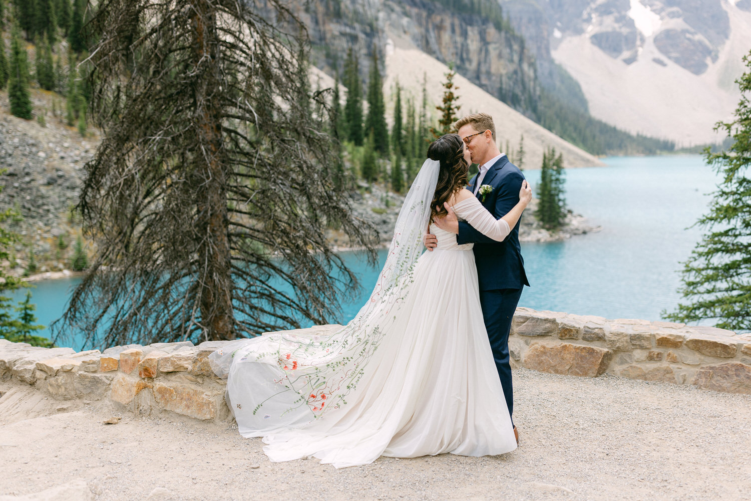 A couple shares a romantic kiss by a stunning blue lake, surrounded by towering trees and mountains, capturing a moment of love and joy on their wedding day.