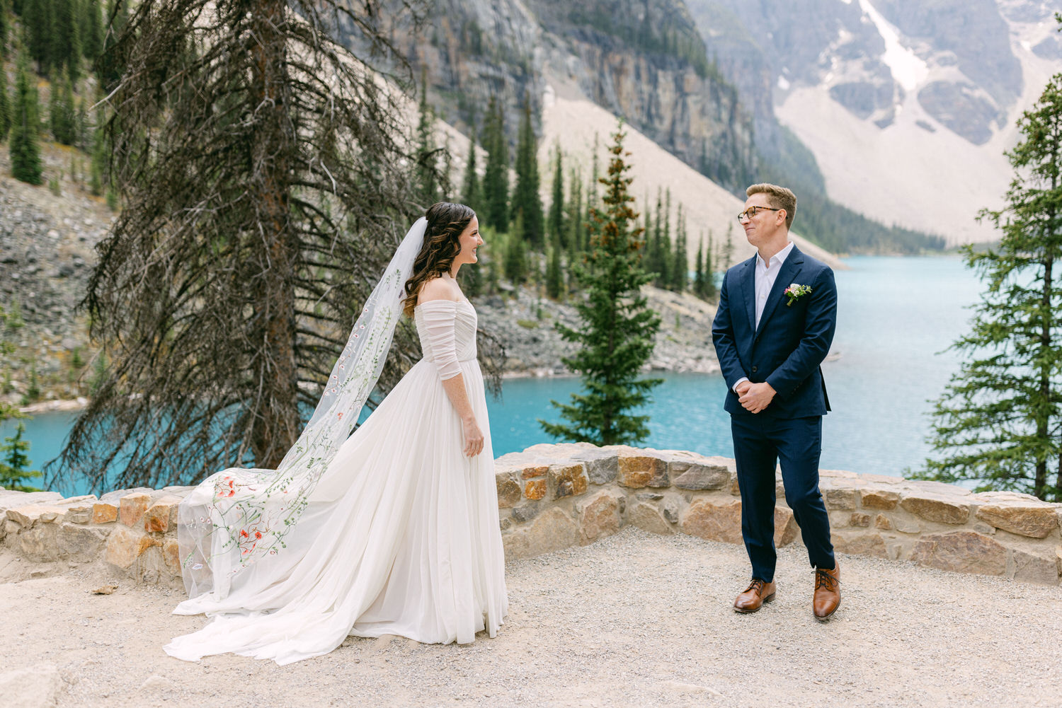 A bride in a flowing white dress and veil stands in front of her groom, who is dressed in a navy suit, with a picturesque turquoise lake and rugged mountains in the background.