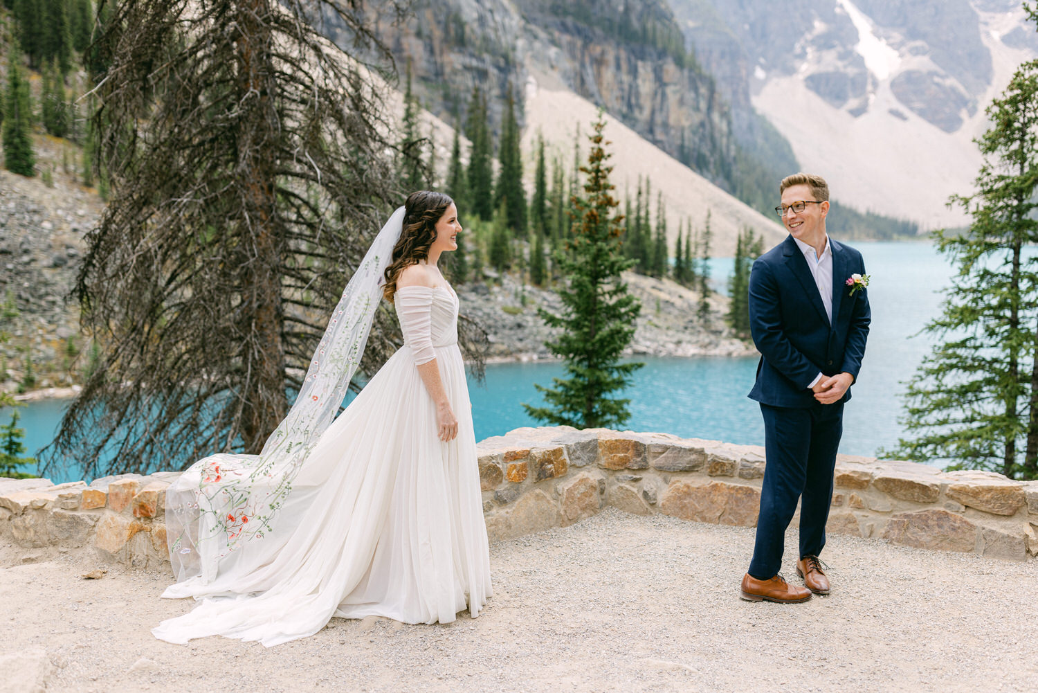 Bride and groom smiling at each other in a picturesque setting by a lake, surrounded by trees and mountains.