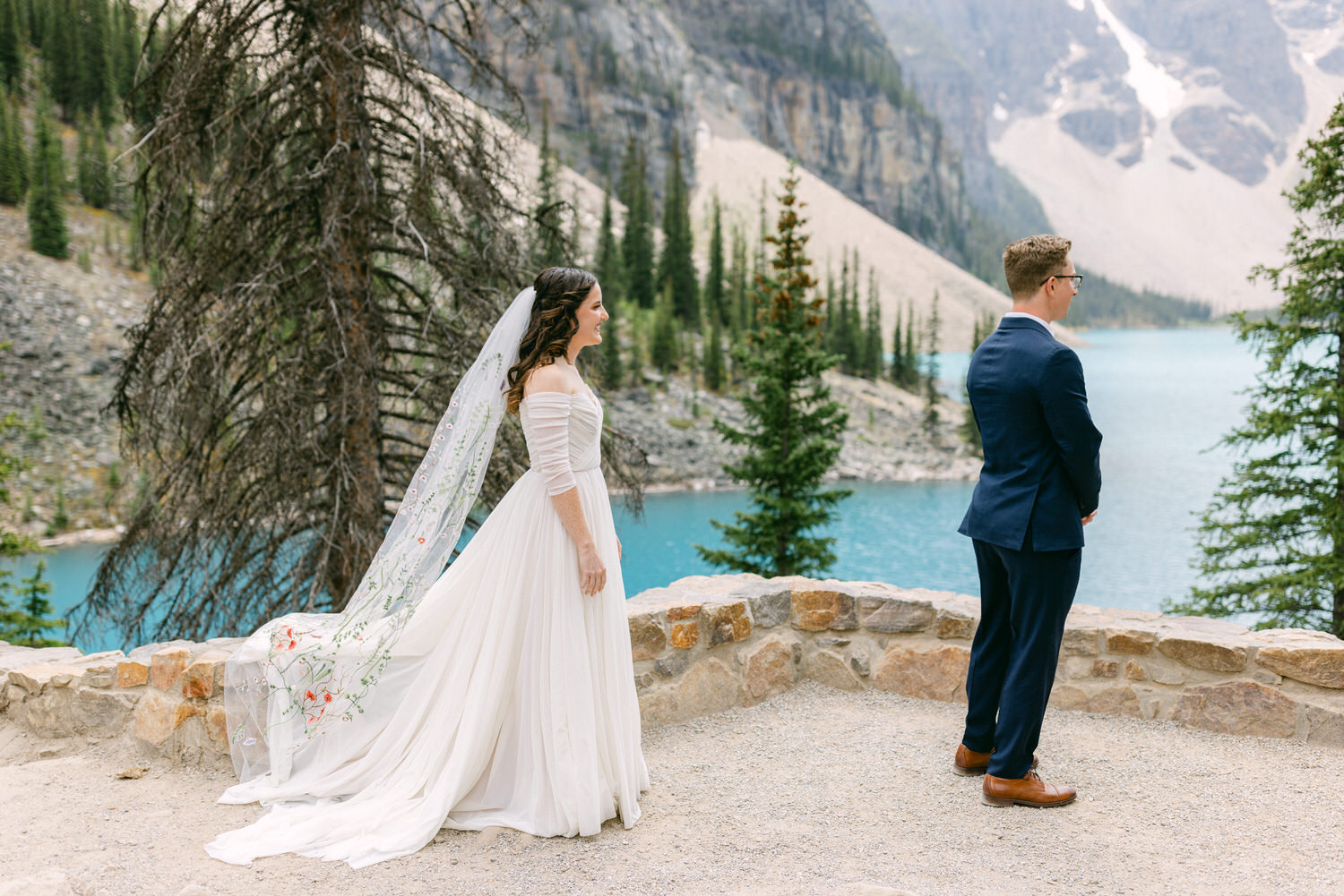 A bride in a flowing white gown and veil stands beside a groom in a blue suit, both gazing at a stunning mountain lake backdrop surrounded by tall trees.