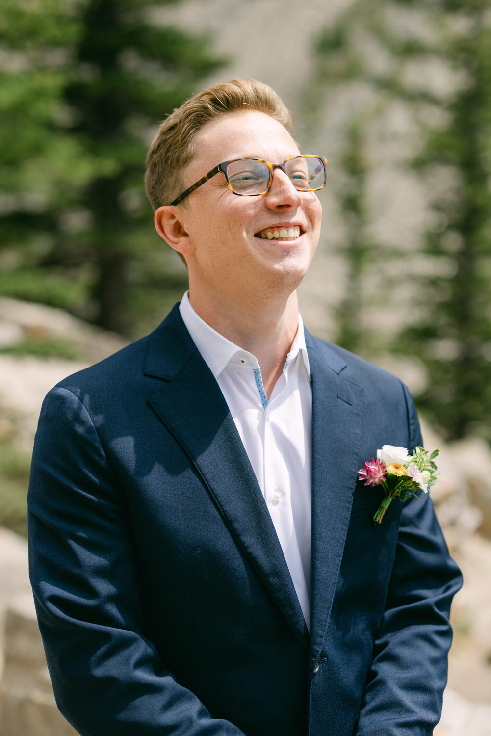 A smiling man in a dark suit and white shirt adorned with a colorful boutonnière, standing outdoors against a backdrop of trees.