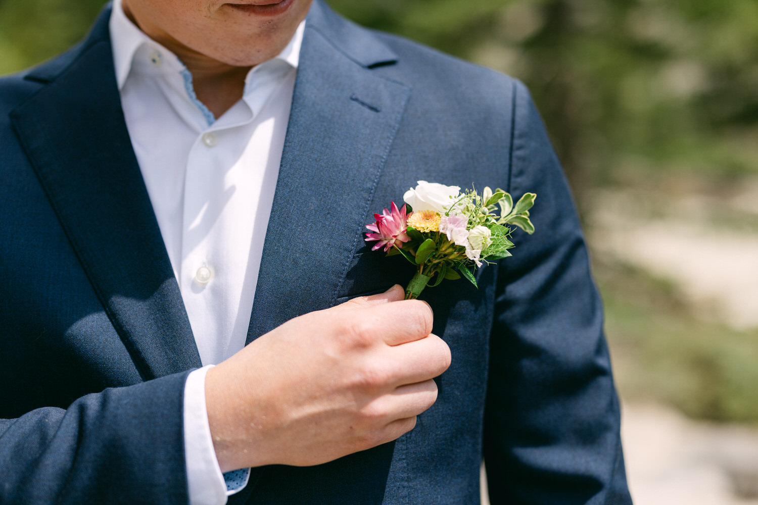 Boutonniere Elegance::A close-up of a man adjusting a colorful boutonniere pinned to his suit jacket, showcasing flowers in shades of pink, white, and green against a backdrop of blurred greenery.