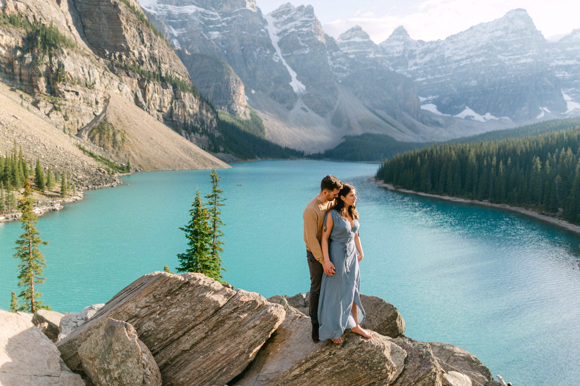 A couple stands on rocks overlooking a turquoise lake surrounded by mountains, with the woman in a flowing blue dress and the man gently embracing her.