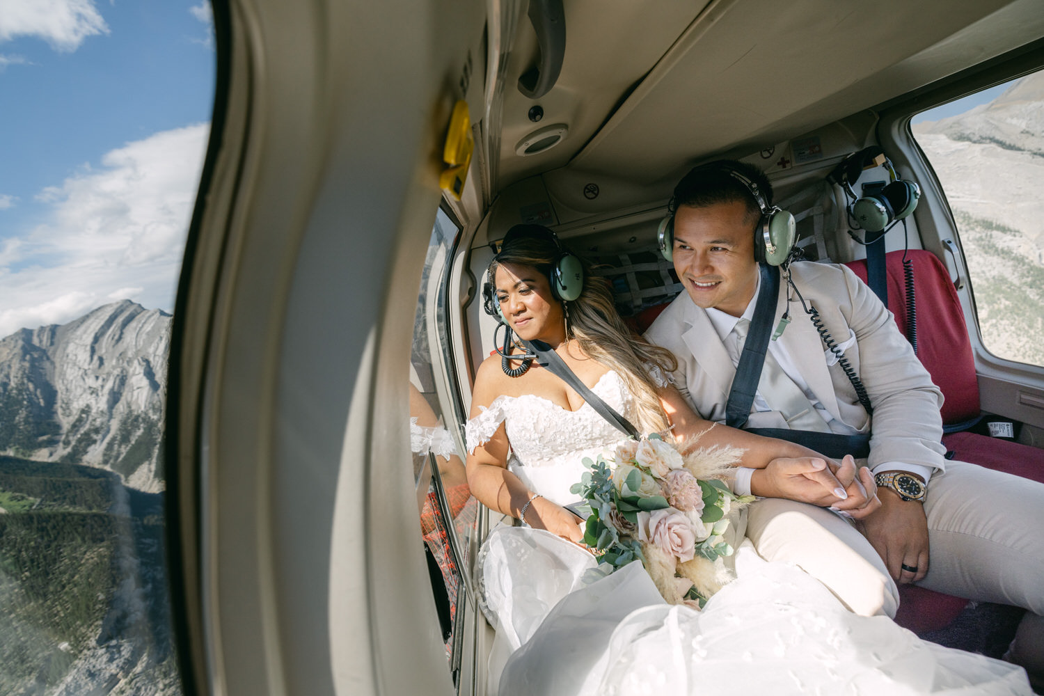 A bride and groom embrace in a helicopter, enjoying stunning mountain views during their unique wedding experience.