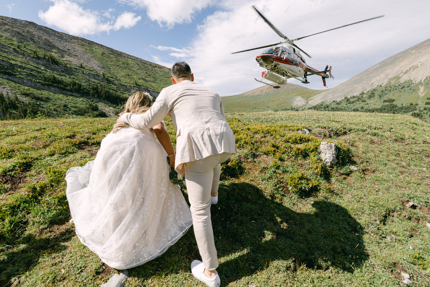 A couple in wedding attire watches a helicopter landing amidst a scenic mountain backdrop.