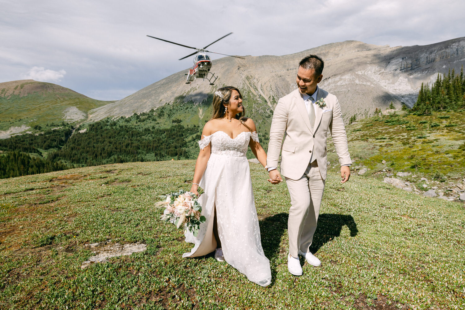 A joyful couple in wedding attire walks hand in hand across a lush green landscape with a helicopter flying above the mountains.