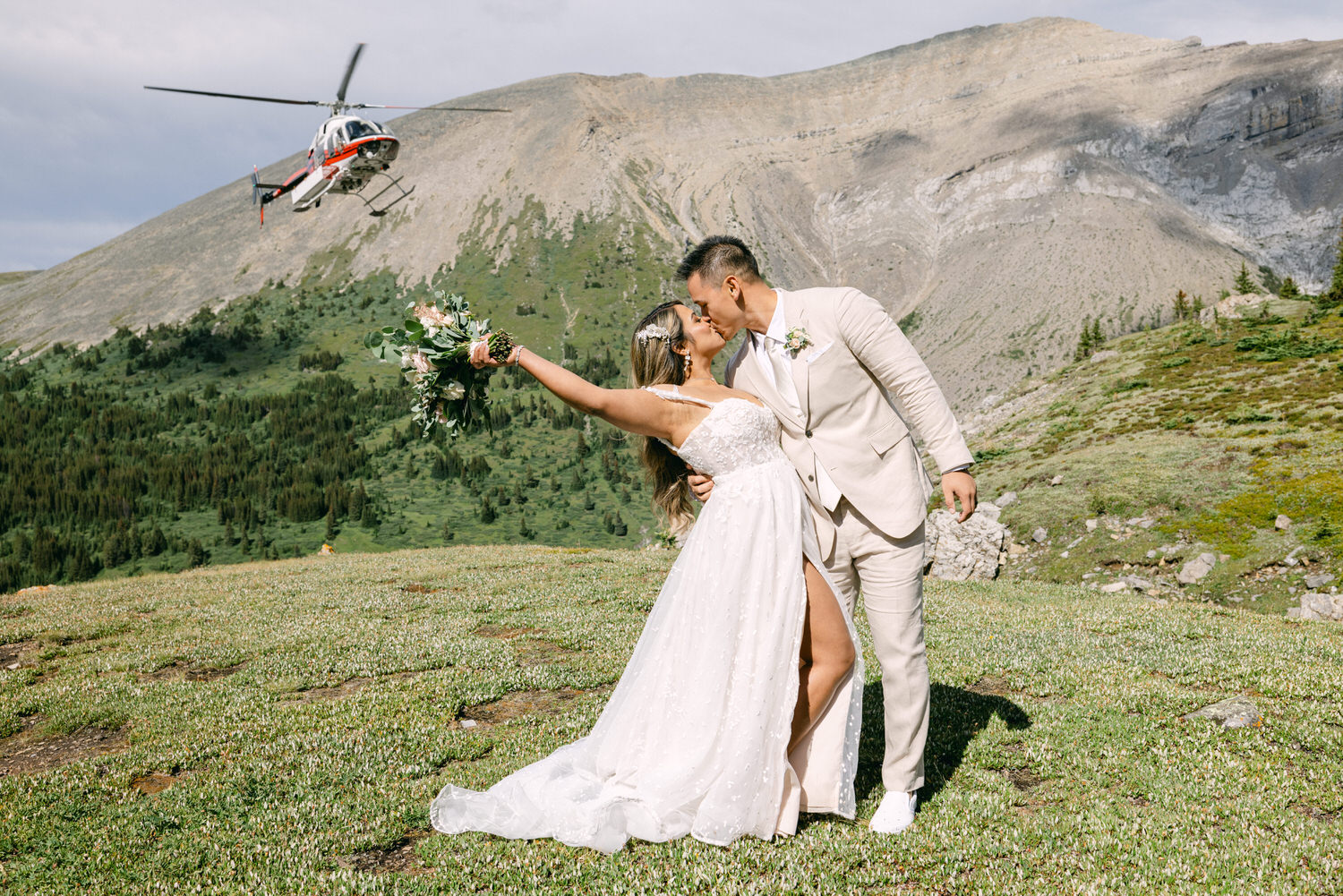 Couple sharing a kiss in a mountainous landscape, with a helicopter flying overhead as the bride holds a bouquet of flowers.