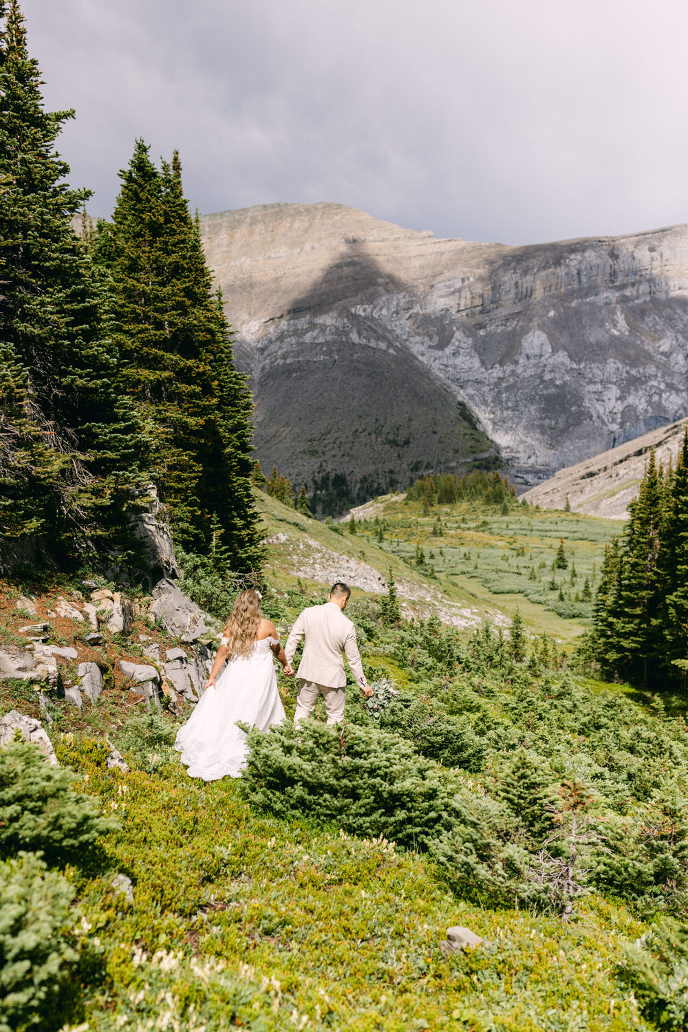 A couple walks hand in hand through a lush mountain landscape, surrounded by trees and rocky terrain, on their wedding day.