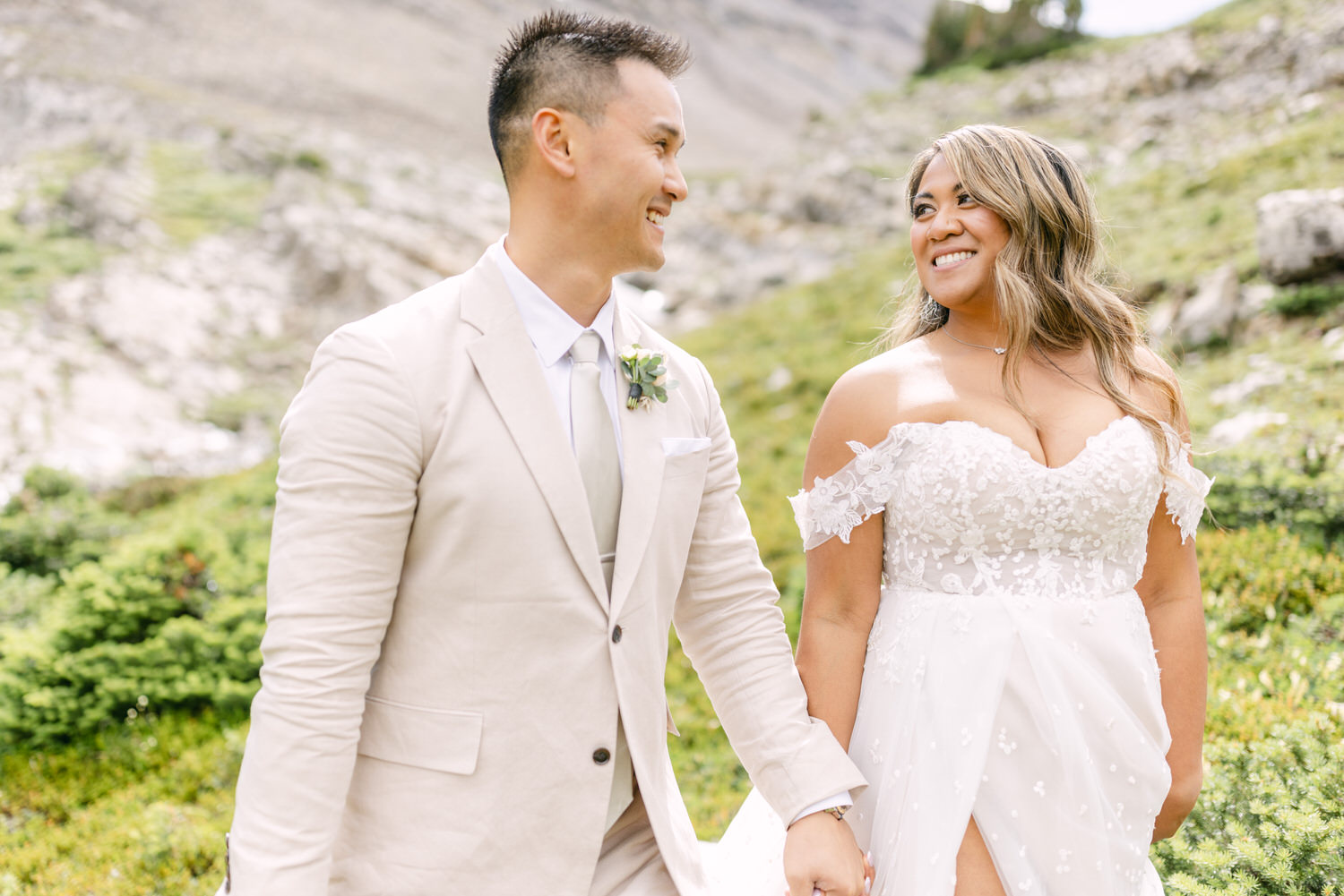 A joyful couple holding hands against a scenic outdoor backdrop, featuring the bride in a stunning lace gown and the groom in a light-colored suit, both beaming with happiness.