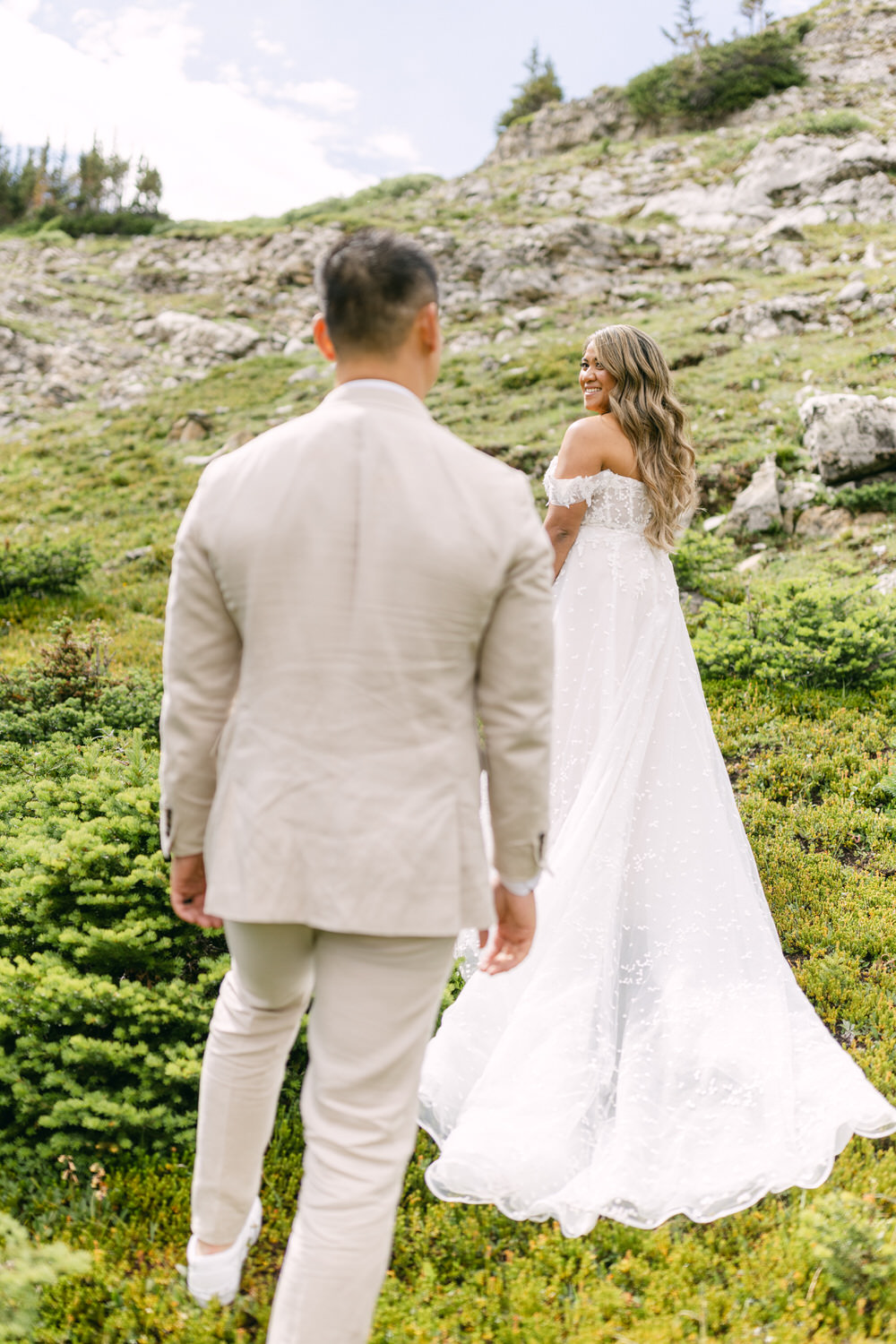 A bride in a flowing white dress smiles back at her groom in a light-colored suit as they stroll through lush greenery on a mountain slope.