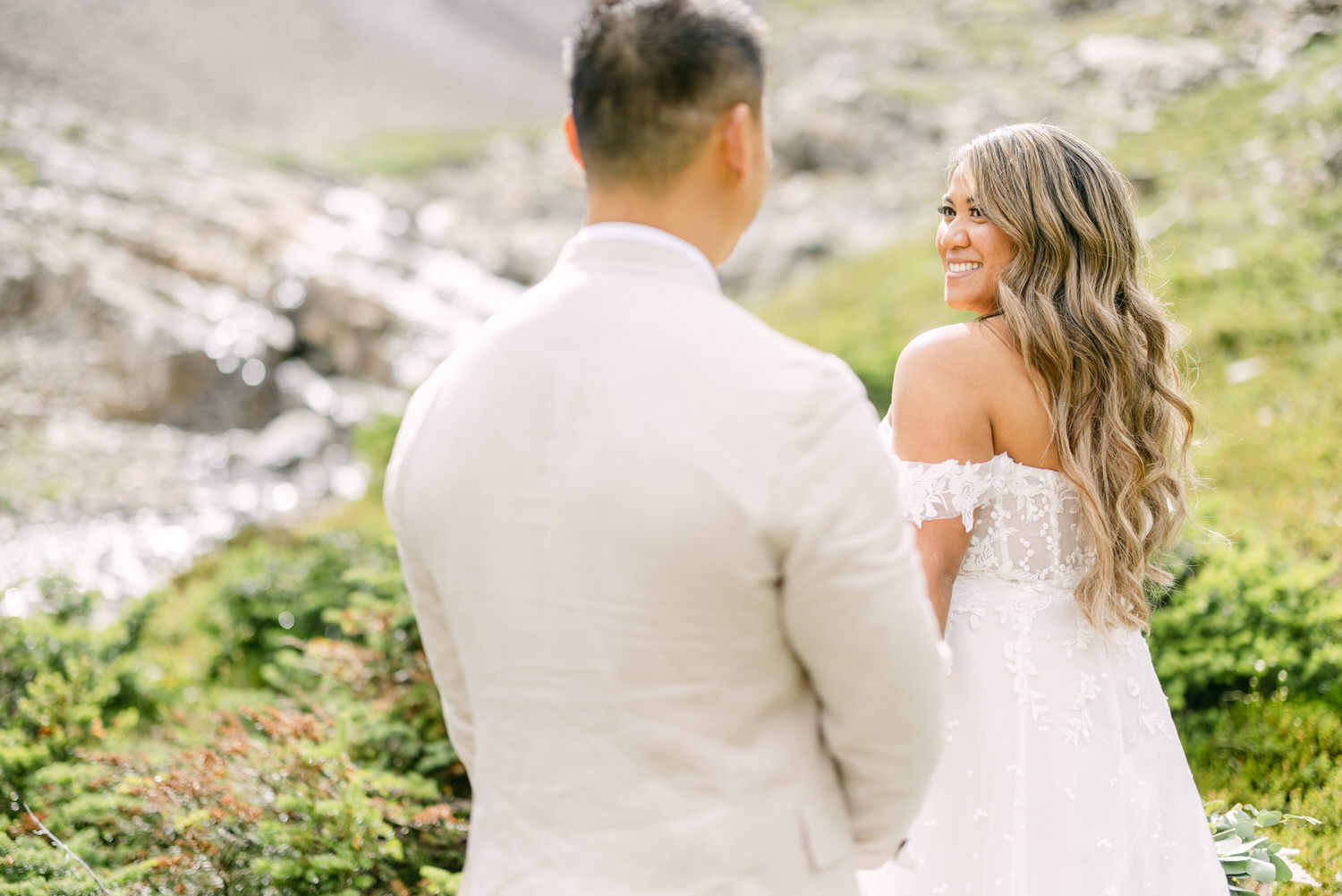 A bride in a floral lace gown smiles at her partner during an intimate moment in a lush, green landscape by a flowing stream.