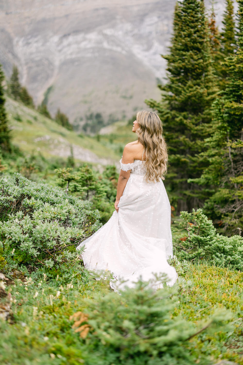 Bride in Nature::A woman in a flowing white dress stands in a lush green landscape, gazing back at the mountains.