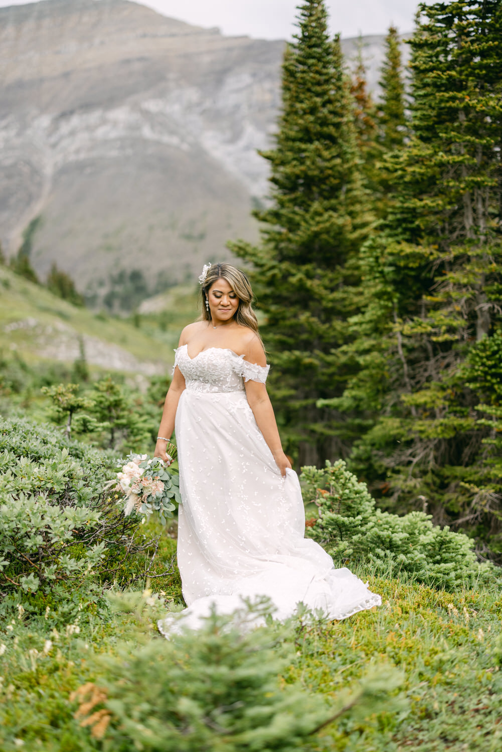 A bride wearing an off-shoulder wedding gown stands gracefully in a lush green setting, holding a bouquet, with a mountain backdrop.