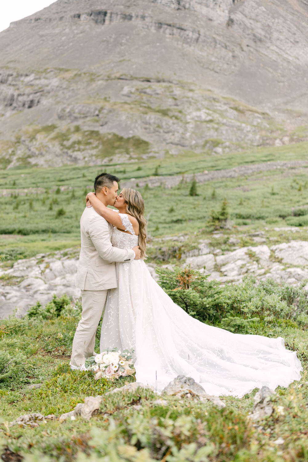 A couple embraces in a picturesque outdoor setting, surrounded by lush greenery and a dramatic mountain backdrop, sharing a tender kiss. The bride wears a flowing white gown, while the groom is dressed in a light suit, highlighting their intimate celebration.