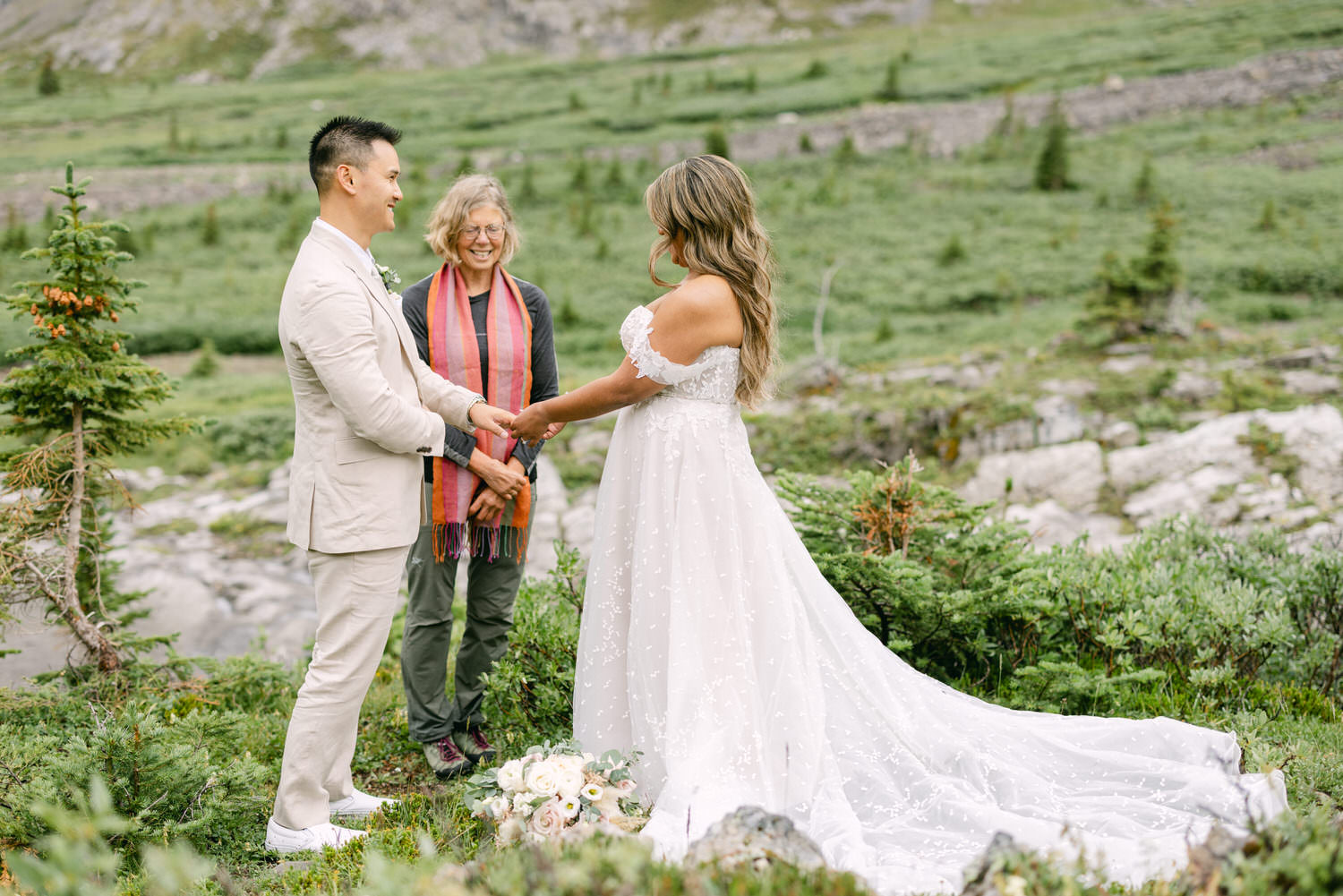 A couple exchanges vows during an outdoor wedding ceremony, surrounded by greenery and a celebrant in a scenic landscape.