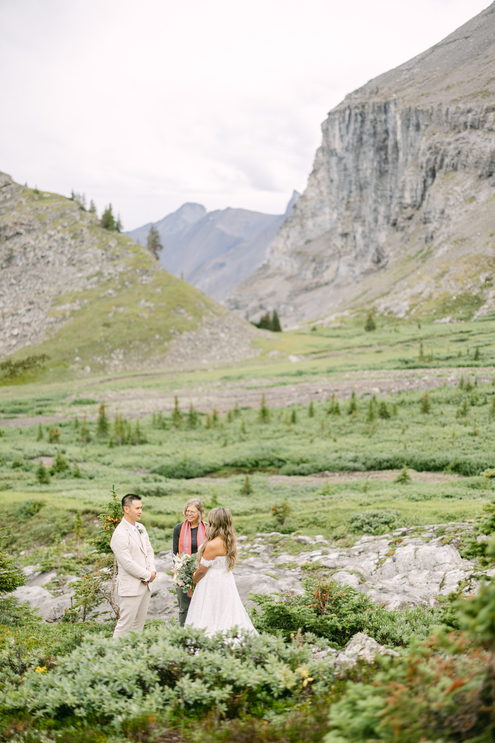 A couple stands together during a wedding ceremony in a picturesque mountainous landscape, surrounded by greenery and a celebrant.