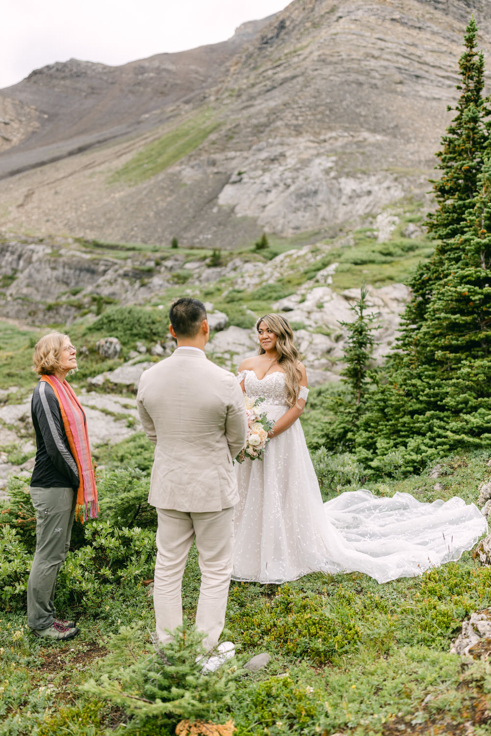 A bride in a flowing white gown holds a bouquet while standing outdoors in a scenic mountain setting, as she faces a groom and an officiant.