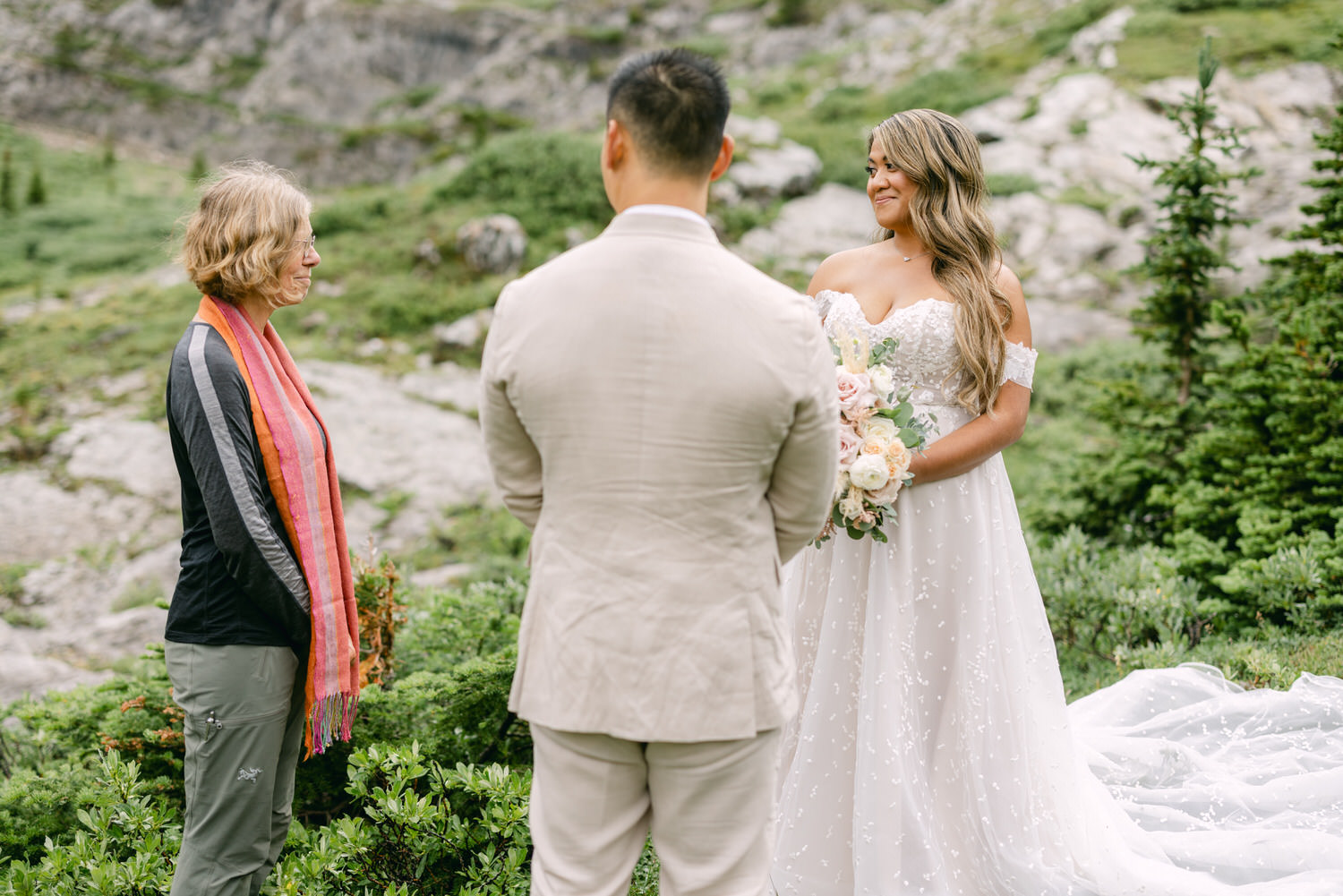 A bride in a flowing white gown with floral details stands with a bouquet, smiling at a man while an officiant in a colorful scarf prepares to conduct the ceremony amidst lush greenery and rocky terrain.