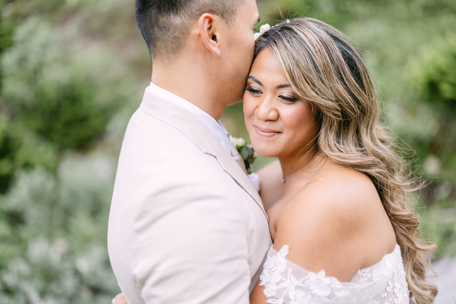 A couple sharing a tender moment, with the groom's forehead touching the bride's, amidst a softly blurred natural background.