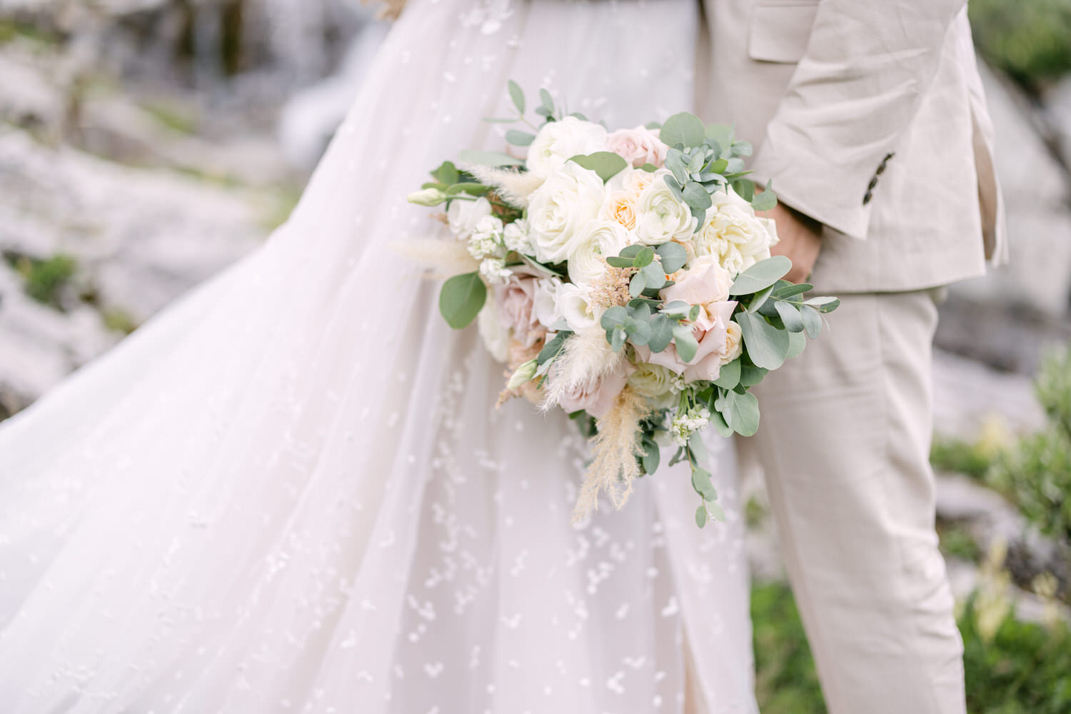 Close-up of a wedding bouquet featuring soft pink and white flowers, surrounded by greenery, held by a couple in a serene outdoor setting.