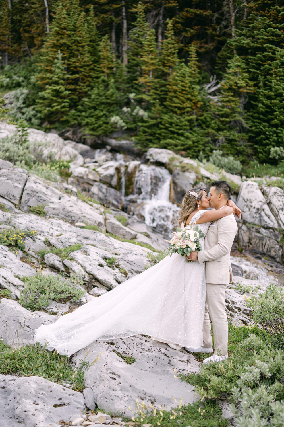 A smiling couple embraces on a rocky landscape near a waterfall, surrounded by lush greenery, with the bride in a flowing white dress and the groom in a light suit.