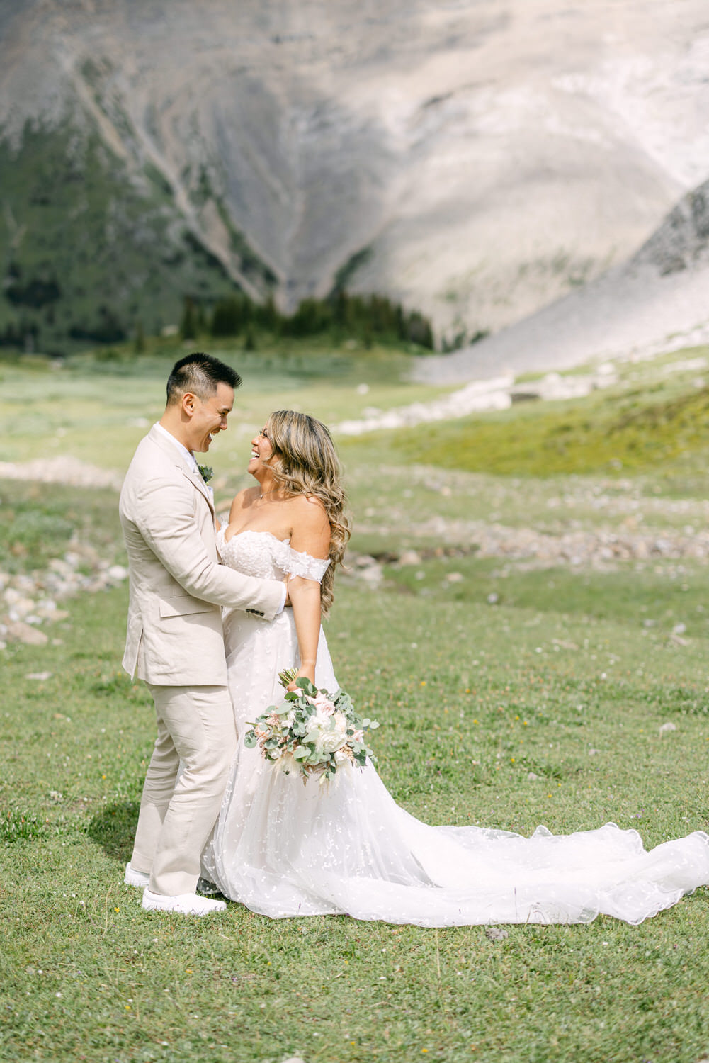 A joyful couple shares a loving moment in a picturesque mountain setting, with the bride in a flowing white gown holding a bouquet and the groom in a light suit.