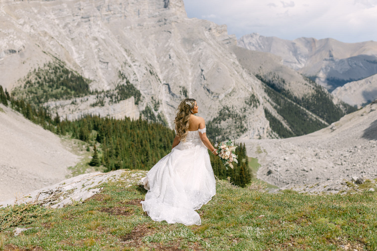 A bride in a flowing white gown holding a bouquet stands on a grassy ledge overlooking a breathtaking mountain landscape.