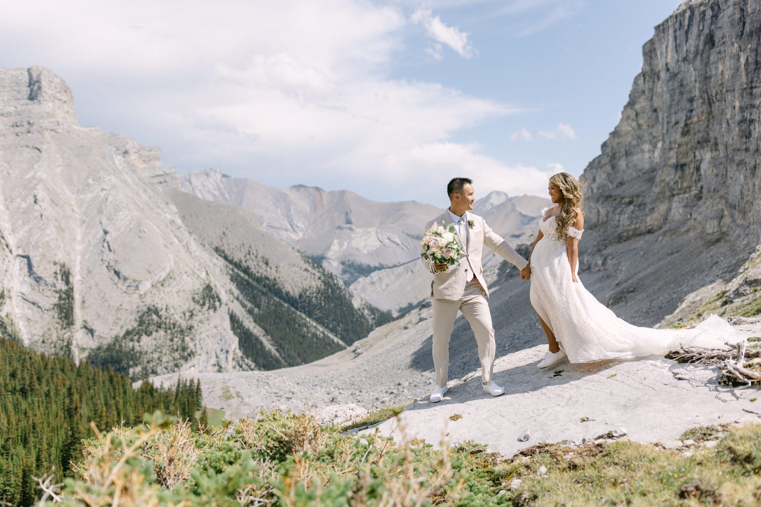 A couple holding hands on a rocky mountain landscape, with the bride in a flowing white gown and the groom in a light suit, surrounded by breathtaking nature.