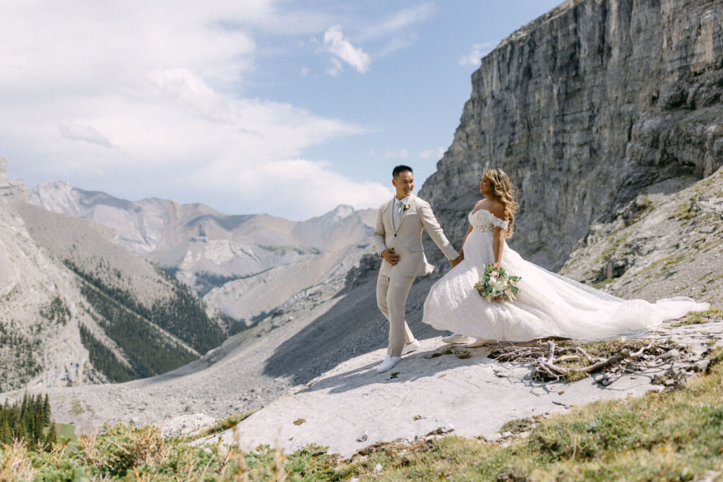 A happy couple strolls hand in hand through a picturesque mountain landscape, showcasing their wedding attire amidst stunning natural scenery.