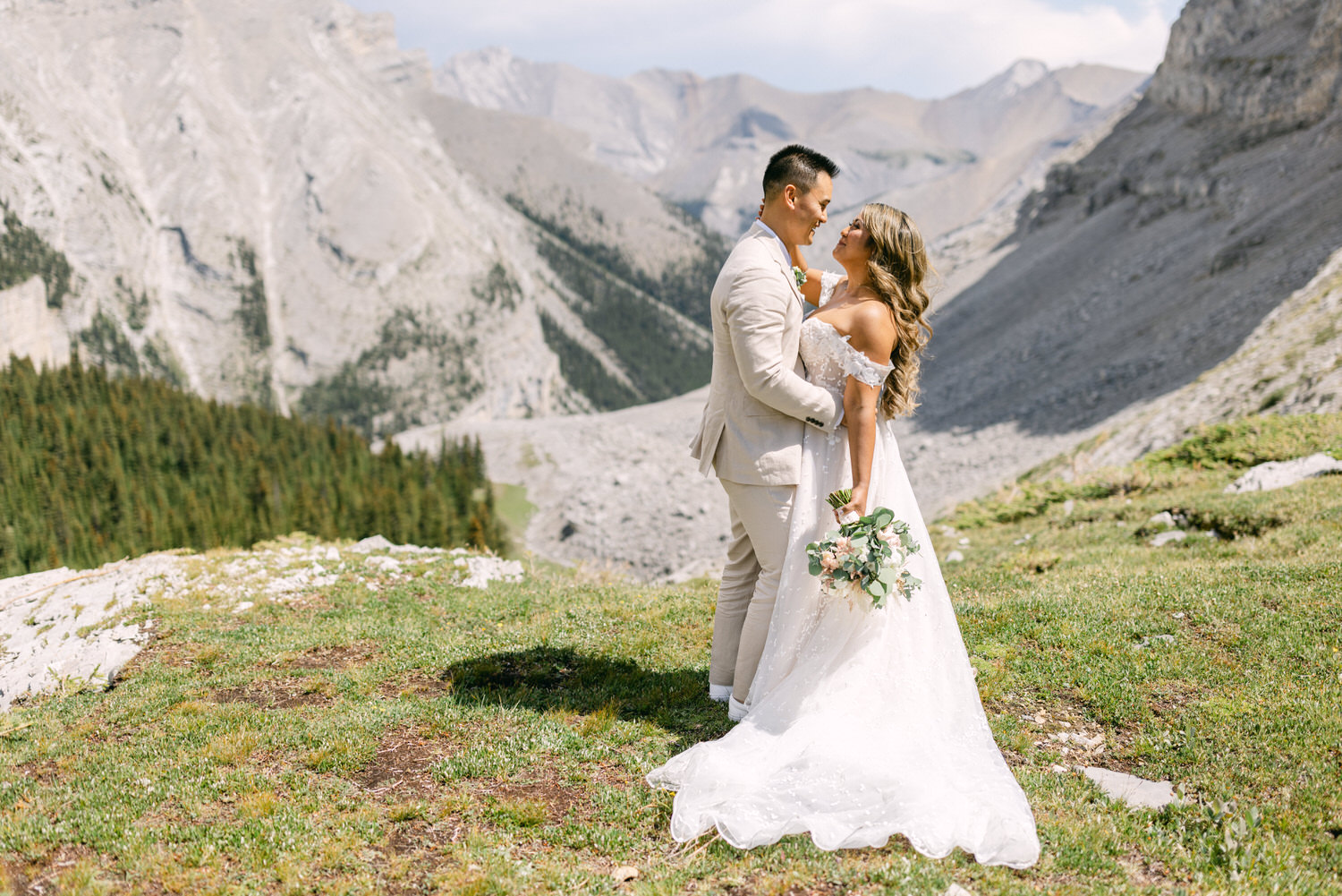 A bride and groom embrace in a picturesque mountain landscape, surrounded by lush greenery and rocky peaks, capturing a romantic moment on their special day.