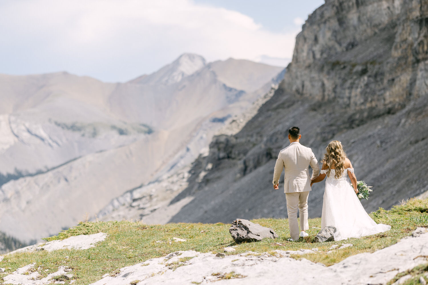 Couple holding hands in bridal attire, standing on a rocky terrain with majestic mountains in the background.