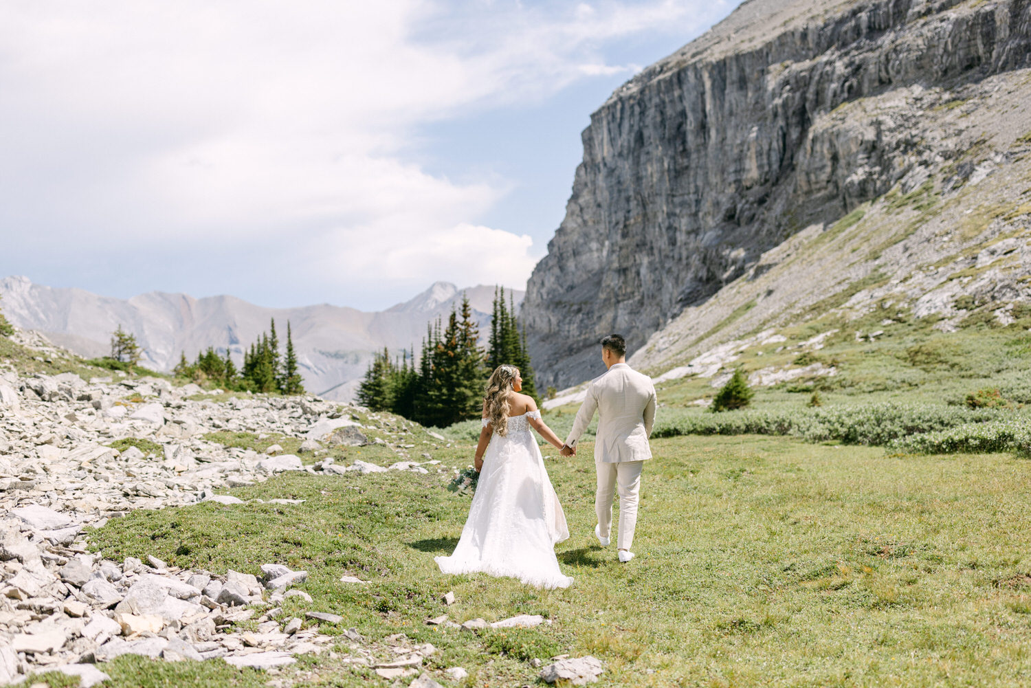 A couple dressed in wedding attire walks hand in hand through a picturesque rocky landscape, surrounded by mountains and greenery.