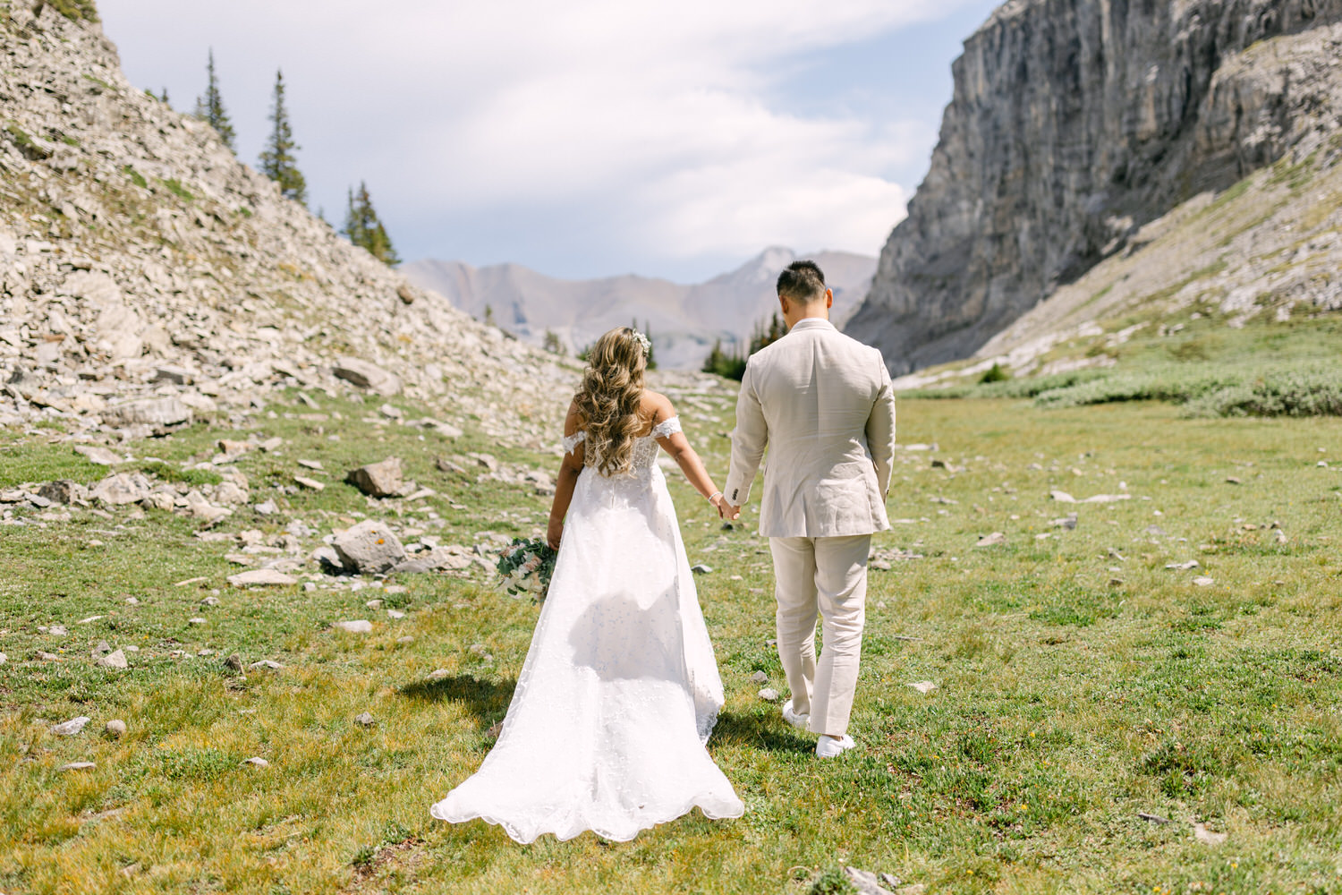 A couple walks hand-in-hand through a picturesque mountain landscape, surrounded by rocky terrain and lush greenery, with the bride's flowing gown and floral bouquet contrasting against the scenery.