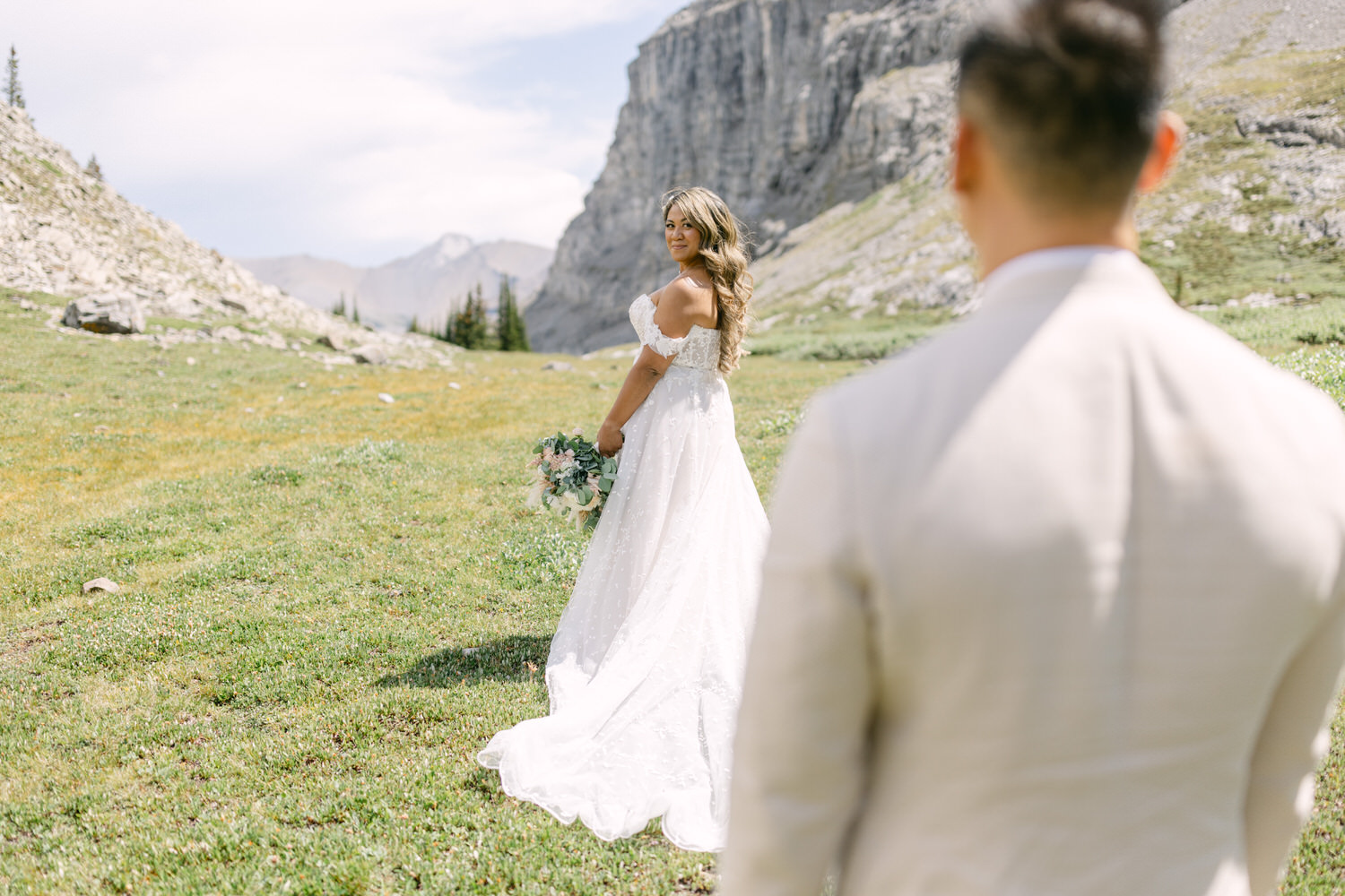 A bride in a flowing white gown stands in a scenic meadow, looking back at her partner, surrounded by mountains and greenery.