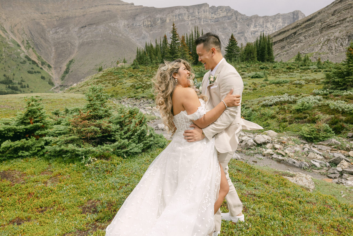 A joyful bride and groom share a loving embrace in a lush, mountainous setting, with vibrant greenery and rocky terrain in the background.