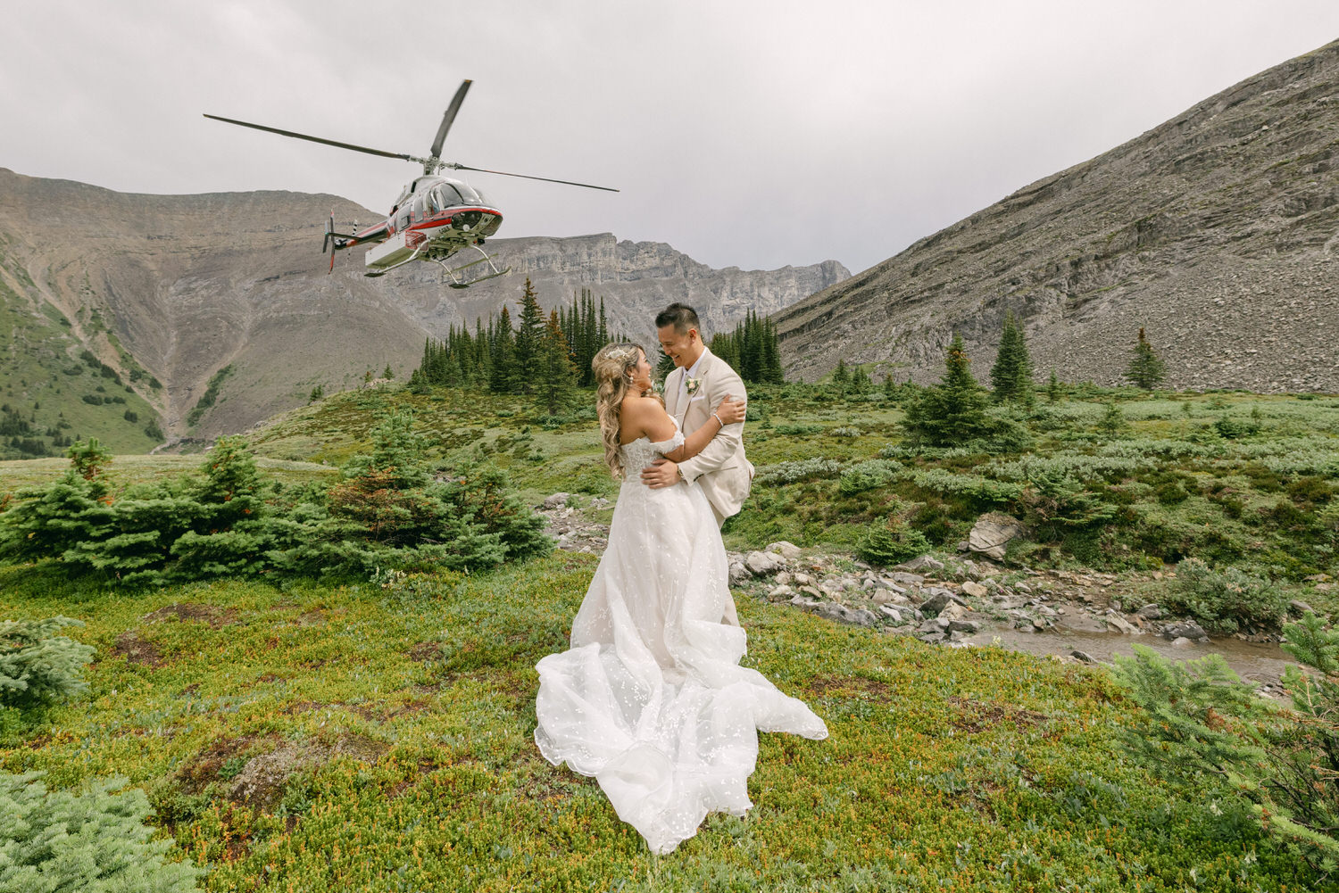 A couple embraces in a lush green landscape with a helicopter flying overhead, capturing a moment of romance amidst nature.