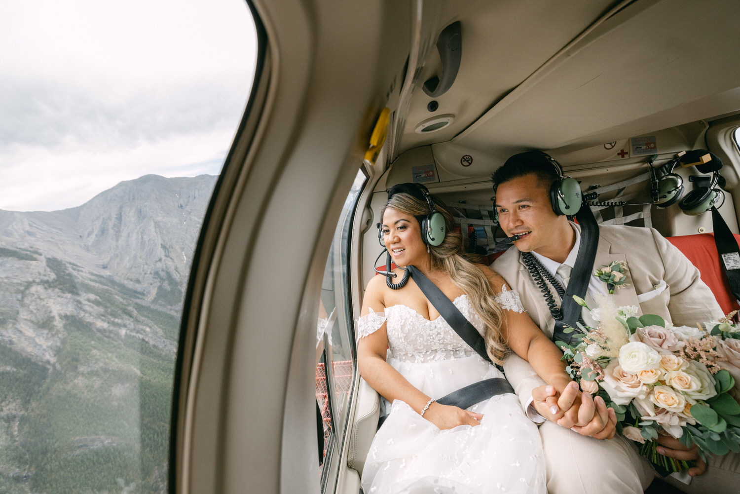 A joyful couple in wedding attire holding hands inside a helicopter, gazing out at mountainous scenery while wearing headsets.