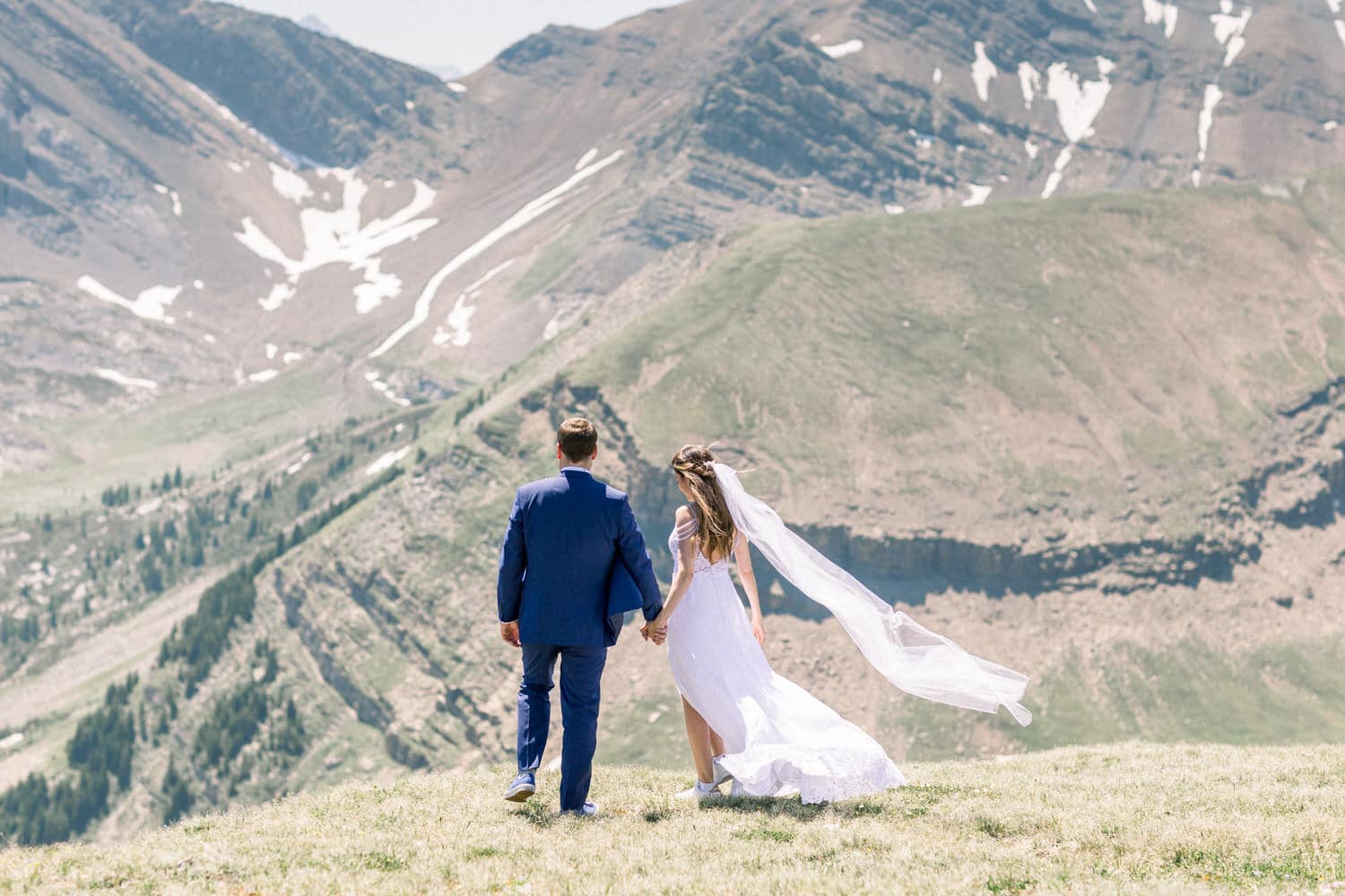 A couple in wedding attire holding hands while walking on a grassy hillside with majestic mountains in the background.